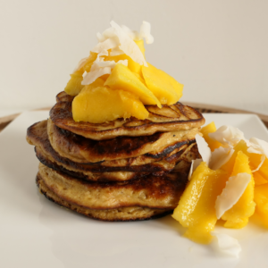 Live Puri Proteïne pancakes met stukken mango en kokossnippers op wit bord met houten ondergrond